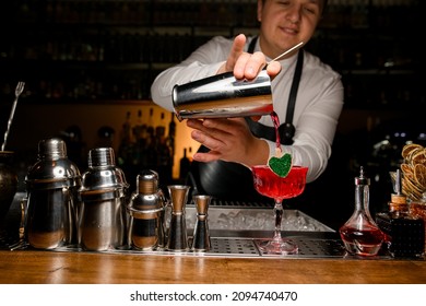 Smiling male bartender neatly pouring red fresh alcoholic drink into glass decorated with small wooden heart on the metallic bar counter. Jiggers and shakers standing near.