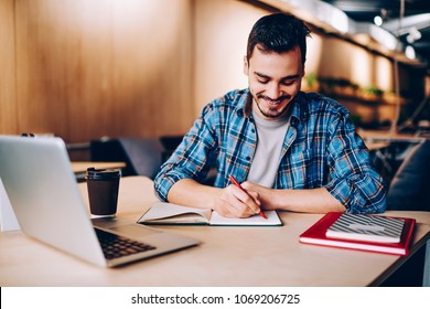 Smiling Male Blogger Satisfied With Having Great Idea For Publication Making Notes Sitting At Desktop With Netbook, Positive Student Enjoying Learning Process Doing Homework Task In College Library