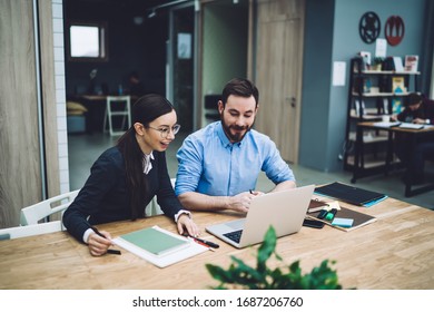 Smiling male in blue shirt with rolled up sleeves sitting near young dark haired female in glasses interested in using laptop at workplace - Shutterstock ID 1687206760