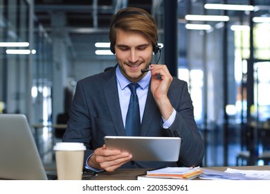 Smiling Male Business Consultant With Headphones Sitting At Modern Office, Video Call Looking At Laptop Screen. Man Customer Service Support Agent Helpline Talking Online Chat