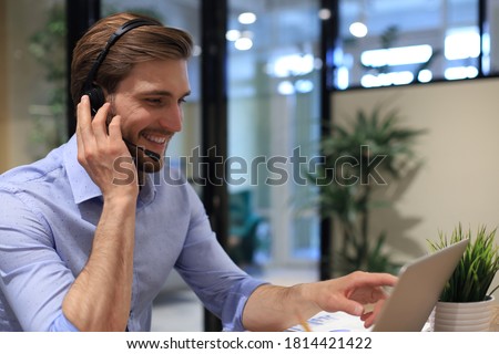 Smiling male business consultant with headphones sitting at modern office, video call looking at laptop screen. Man customer service support agent helpline talking online chat.