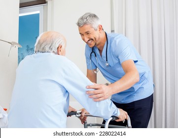 Smiling Male Caretaker Helping Senior Man To Use Walking Frame In Bedroom At Nursing Home