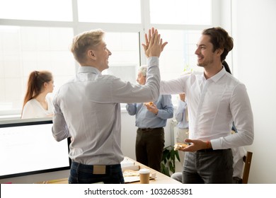 Smiling Male Coworkers Giving High Five Eating Pizza Together At Office Break, Happy Young Men Colleagues Slapping Hands Celebrating Success Achievement At Staff Party, Teammates Friendship Concept