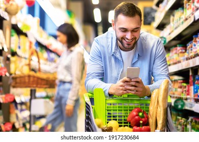 Smiling Male Customer Doing Grocery Shopping Using Smartphone Walking With Cart In Supermarket. Selective Focus. Man Using Groceries Shopping Application On Phone Bying Food In Super Market