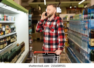 Smiling Male Customer Shopping In A Grocery Store, Talking Using A Smartphone, Walking With A Trolley In A Supermarket. A Man Using A Grocery Shopping App On His Phone Buys Food In A Supermarket