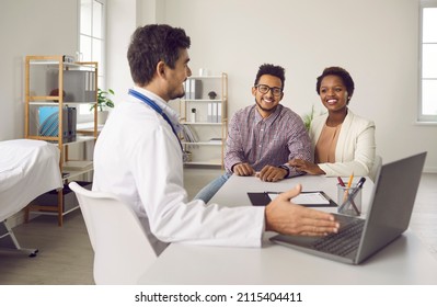 Smiling Male Doctor Consult Excited Multiethnic Family Clients Show Results On Computer In Clinic. Man GP Or Gynecologist Have Meeting With Ethnic Couple For Pregnancy Planning In Hospital.