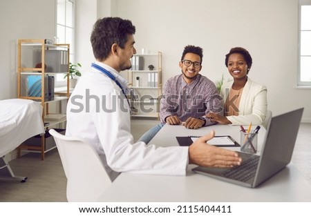 Smiling male doctor consult excited multiethnic family clients show results on computer in clinic. Man GP or gynecologist have meeting with ethnic couple for pregnancy planning in hospital.