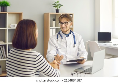 Smiling male doctor discussing treatment with female patient. Young physician in white lab coat with stethoscope talking to woman sitting at office desk with notebook PC in modern clinic or hospital