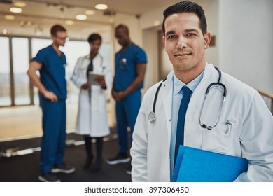 Smiling male doctor wearing white jacket and stethoscope with three diverse colleagues in background