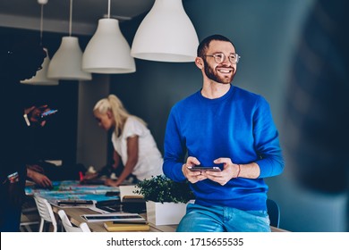 Smiling male employee in casual clothes and eyeglasses sitting on table in modern workplace with diverse colleagues and browsing tablet while resting during work and looking away