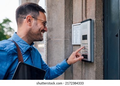 Smiling Male Executive Using Intercom By Door. Young Businessman Is Holding Digital Tablet. He Is Standing At Office Building Entrance In The City.