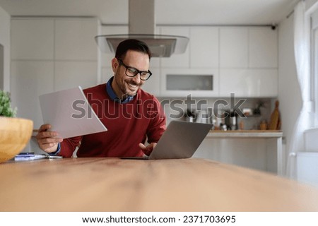 Smiling male financial adviser holding reports and working over laptop at desk in home office