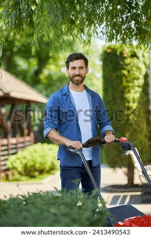 Smiling male gardener in denim shirt operating modern lawn mower outdoors. Portrait of happy man posing with grass cutter, looking at camera, while working in garden in summer. Concept of gardening. 