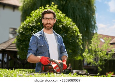 Smiling male landscaper in safety glasses cutting bush with hedge trimmer on backyard. Portrait of happy bearded gardener pruning boxwood with bush cutter and looking at camera. Concept of workplace. - Powered by Shutterstock