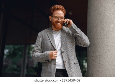 Smiling male manager in eyeglasses is talking phone standing near office building - Shutterstock ID 2395459409