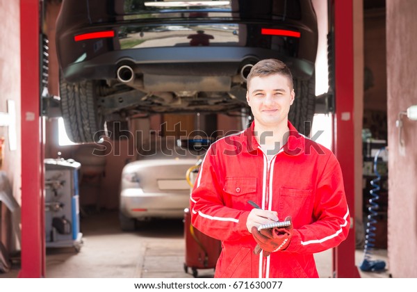 Smiling male motor mechanic standing making\
notes in front of a black sedan elevated on a hoist in a bay in a\
garage or workshop