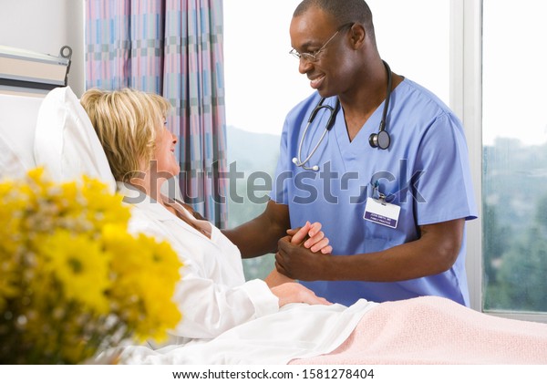 Smiling male nurse holding hands with female patient\
in hospital bed