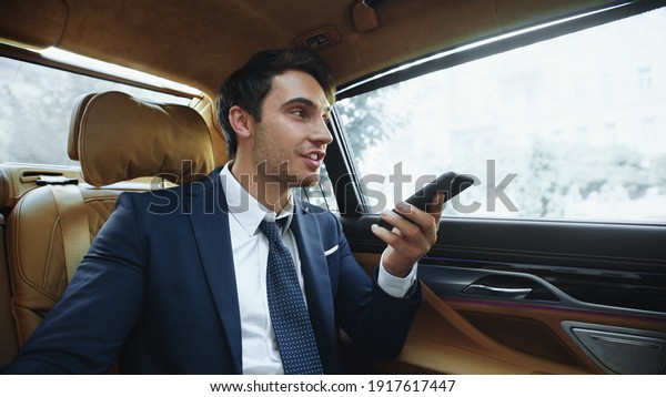 Smiling male professional recording voice\
message to smartphone in business car. Friendly business man\
working with cellphone in modern car. Handsome man dictating\
message on phone in\
automobile.