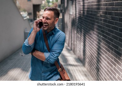 Smiling Male Professional Talking On Smart Phone. Happy Businessman Is Carrying Laptop And Business Bag. He Is Walking On Footpath At City During Sunny Day.