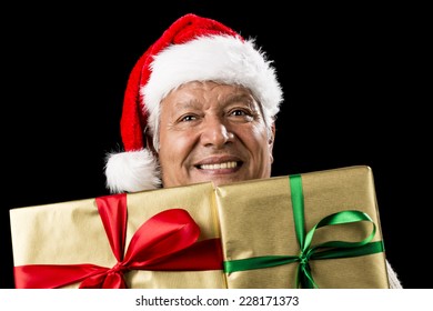 Smiling Male Senior Peeping Across Two Foot-long Gifts That Cover His Chest And Chin. Both Presents Wrapped Golden With Green And Red Bows. Father Christmas Hat. Gift Giving Theme Isolated On Black.