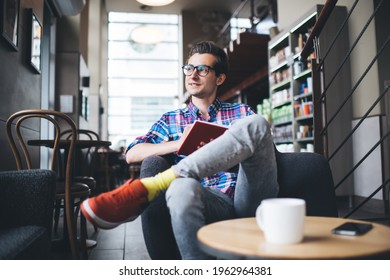 Smiling male student in casual wear and eyeglasses sitting in armchair with legs crossed and thinking looking away while spending free time in cozy cafe with cup of coffee and notebook in hands - Powered by Shutterstock