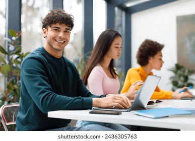 Smiling male student with curly hair sitting with diverse female classmates in sunny, plant-filled study space, all focused on their individual laptops - Powered by Shutterstock