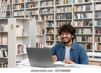 Smiling Male Student Studying Online Watching Webinar Using Laptop Having Video Call With Teacher In University Library With Bookshelf On Background. Remote Education Course, Elearning