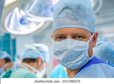 Smiling male surgeon in uniform with mask in surgery room  with assistent