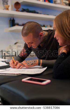 Smiling male tattooist and blonde girl creating sketch for tattoo.