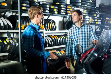 Smiling male technician standing with customer in shop with motorcycles. Focus on the right man
