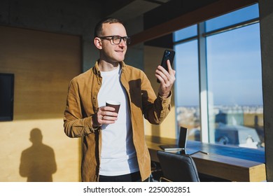 Smiling male worker in casual clothes looking away while talking on mobile phone and drinking coffee during break in modern creative workspace - Shutterstock ID 2250032851