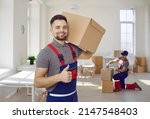 Smiling male worker of moving and delivery company holding cardboard box showing thumbs up. Loader in overalls posing against background of colleague who packs cardboard boxes. Moving service concept.