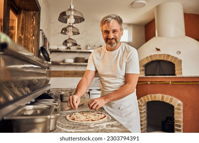 Smiling male worker, working at the pizzeria, making pizzas.