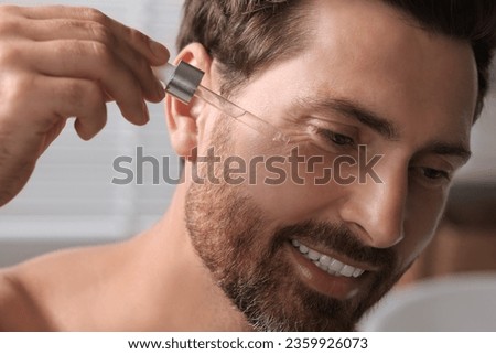Smiling man applying cosmetic serum onto his face indoors, closeup