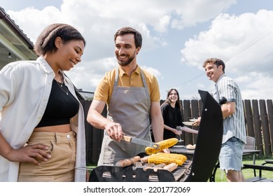 Smiling Man In Apron Cooking Corn On Grill Near Bi-racial Friend In Backyard