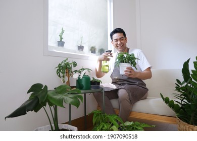 Smiling man Asian in apron sitting on cozy sofa watering plant  in green house. Best Hobby in weekend. - Powered by Shutterstock