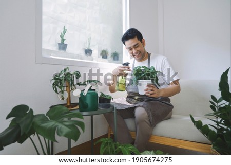 Smiling man Asian in apron sitting on cozy sofa watering plant  in green house. Best Hobby in weekend.