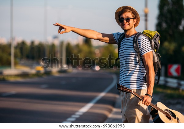 smiling man with backpack and guitar in hand\
gesturing to stop car while\
hitchhiking