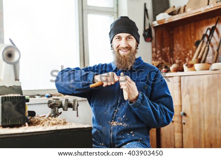 Smiling man with a beard wearing blue jeans suit and black hat carving wooden spoon with instruments and looking at camera, woodcarving instruments on shelf.