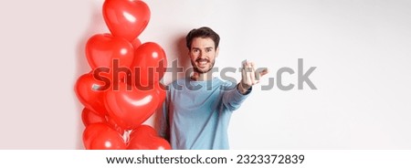 Smiling man beckon you to come closer, follow me gesture, taunting his lover move forward, have romantic surprise, standing near red balloon on valentines day, white background.