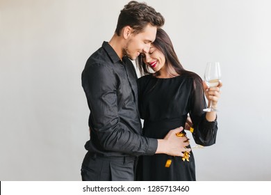 Smiling Man In Black Outfit Holding Golden Confetti And Dancing With Shy Brunette Girl. Pretty Dark-haired Woman Relaxing With Husband At Party And Drinking Wine.