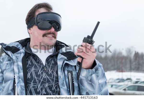 Smiling man in black ski goggles holds radio\
standing outdoor.