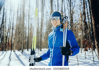 Smiling man in a blue jacket, holding skis and poles, prepares for cross-country skiing on a sunny winter day in a snowy forest.  - Powered by Shutterstock