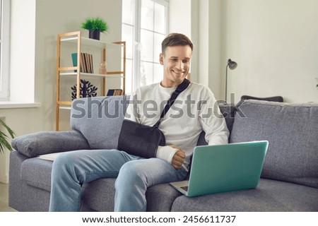 Smiling man with a broken arm in consequence injury, swathed in a protective cast and bandage, showcases resilience as he comfortably despite the trauma uses a laptop on the living room sofa at home.