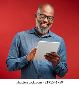 Smiling Man Browsing On A Tablet Online, Networking On The Internet And Scrolling On An App While Standing Against A Red Studio Background. Happy, Mature And African Male Reading An Email And Typing