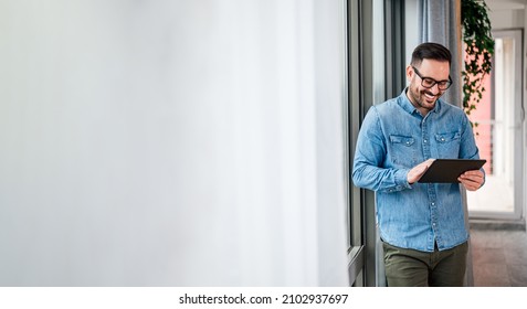 Smiling Man Businessman In Casuals Standing In Office Next To Window Using Tablet Making Online Shopping Purchasing Small Business Home Office Entrepreneur Looking At Tablet Making Online Video Call