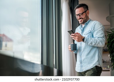 Smiling Man Businessman In Casuals Standing In Office Next To Window Using Mobile Phone Drinking Coffee Holding Coffee Cup Small Business Entrepreneur Looking At Smart Phone While Taking Coffee Break