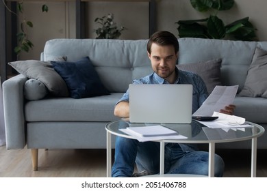 Smiling Man Calculating Domestic Bills, Taxes, Using Laptop, Browsing Online Banking Service, Sitting On Floor In Living Room, Young Tenant Renter Or Freelancer Checking Financial Documents