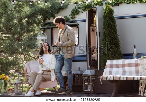 Smiling man in cardigan standing near girlfriend with\
wine and camper van