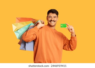 Smiling man carrying colorful shopping bags and showing a credit card, on a plain yellow background - Powered by Shutterstock